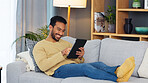 Young man happily laying on couch with tablet. Male is relaxing indoors playing games or using social media for fun. He is isolate and smiling full of joy using digital technology for fun