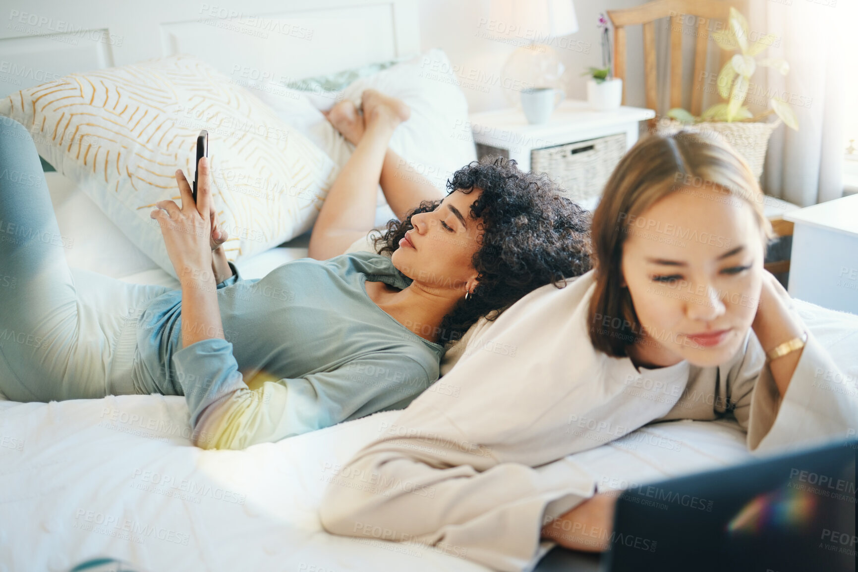 Buy stock photo Lesbian, couple and bed with laptop and phone for streaming movie or internet for relax, connection or online. Lgbt woman, reading and lens flare home for social media or subscription in bedroom