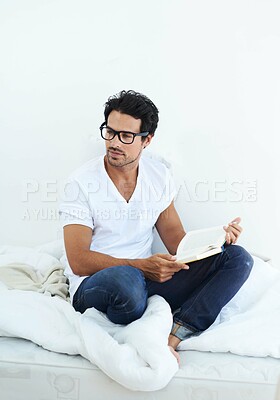 Buy stock photo Shot of a handsome young man sitting on his bed reading a book
