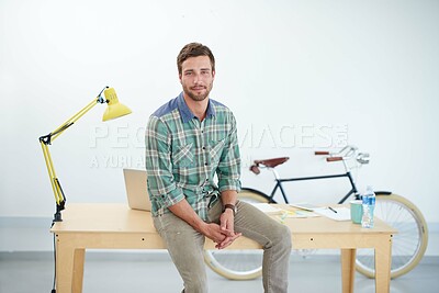 Buy stock photo Portrait of a casually-dressed young man sitting on his desk in an office. The commercial designs displayed represent a simulation of a real product and have been changed or altered enough by our team of retouching and design specialists so that they don't have any copyright infringements