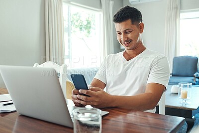 Buy stock photo Cropped shot of a handsome young man using his cellphone while working on a laptop in the living room at home