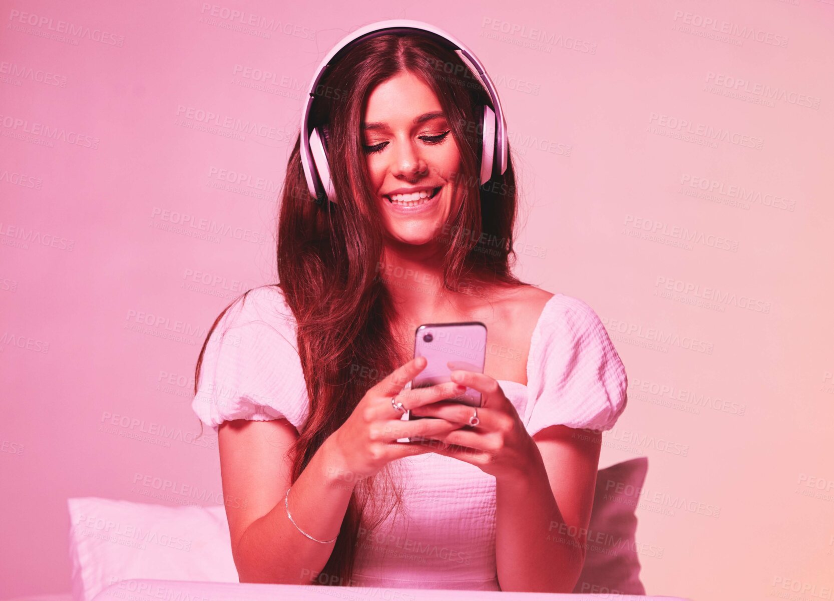 Buy stock photo Pink, light and music by woman in a bed with headphones, social media and streaming on wall background. Podcast, texting and girl relax in bedroom, happy and enjoying radio, audio or online playlist