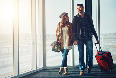 Buy stock photo Shot of a young couple walking inside of an airport with their luggage and holding hands