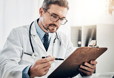 Buy stock photo Shot of a focussed doctor looking at paperwork on a clipboard while sitting at his desk