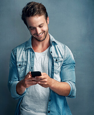 Buy stock photo Shot of a happy young man using his smartphone while standing against a gray background in the studio