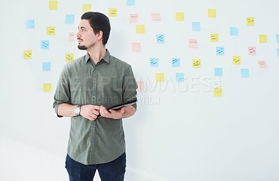 Buy stock photo Shot of a young businessman using a digital tablet while standing against a wall with notes in an office