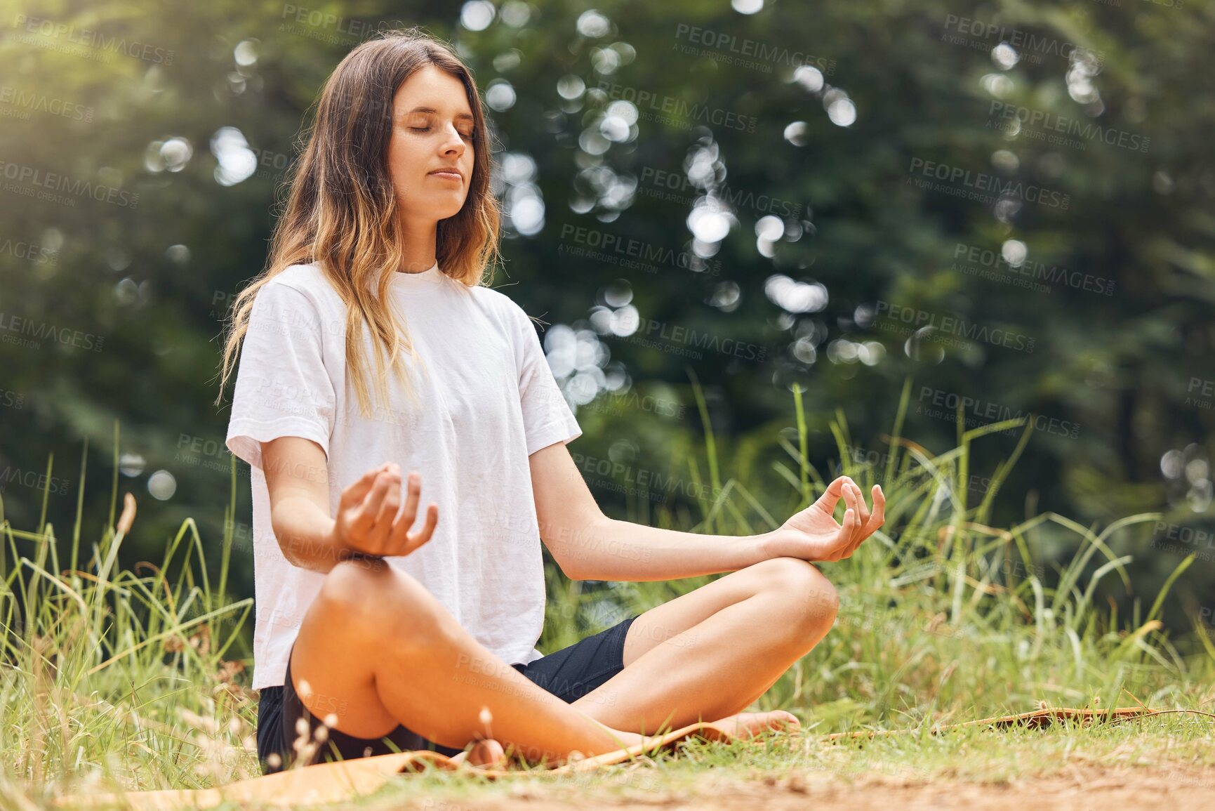 Buy stock photo A calm young caucasian female sitting with her legs crossed doing yoga and meditating on grass in a park on a sunny day. Young woman focusing her zen and energy while working on her mental wellness and relaxing in a forest 




