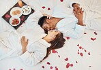 Young couple love on bed, with flower petals and healthy snack on tray while on holiday. Man and woman, romance together in bedroom with roses and food on sheet, smile on romantic trip or vacation.