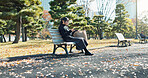 Japanese woman, bench and smartphone in park with relax, happiness and social media in sunshine in city. Female person, sitting and wellness with cellphone in town, streaming and digital technology