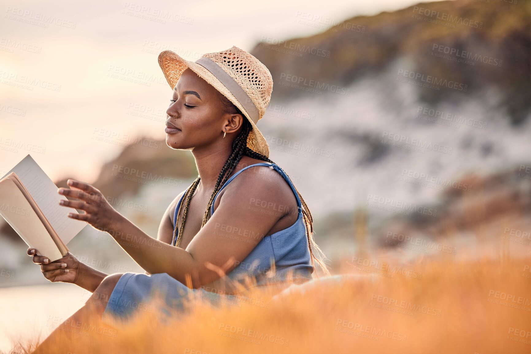 Buy stock photo Mountain, cliff and woman reading a story while relaxing outdoor on the grass at sunset in summer. Freedom, travel and calm African female person enjoying a novel or fantasy book on hill with peace.