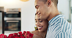 Happy couple, red roses and kiss for surprise, anniversary or valentines day in kitchen at home. Face of young man and woman smile with flowers for romantic gift, love or care in celebration at house