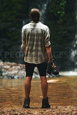 Buy stock photo Shot of a young man staring at a waterfall with his back turned and holding his camera