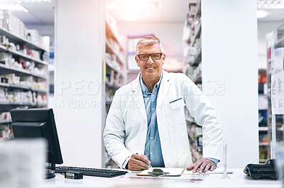 Buy stock photo Portrait of a cheerful mature male pharmacist making notes in a book on the counter while looking at the camera