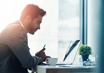 Buy stock photo Shot of a handsome young businessman using a laptop and cellphone at his desk in a modern office