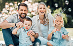 Smiling caucasian family blowing soap bubbles for fun while relaxing together in the park or garden outside on a sunny day. Loving parents bonding with playful kids enjoying a carefree happy childhood