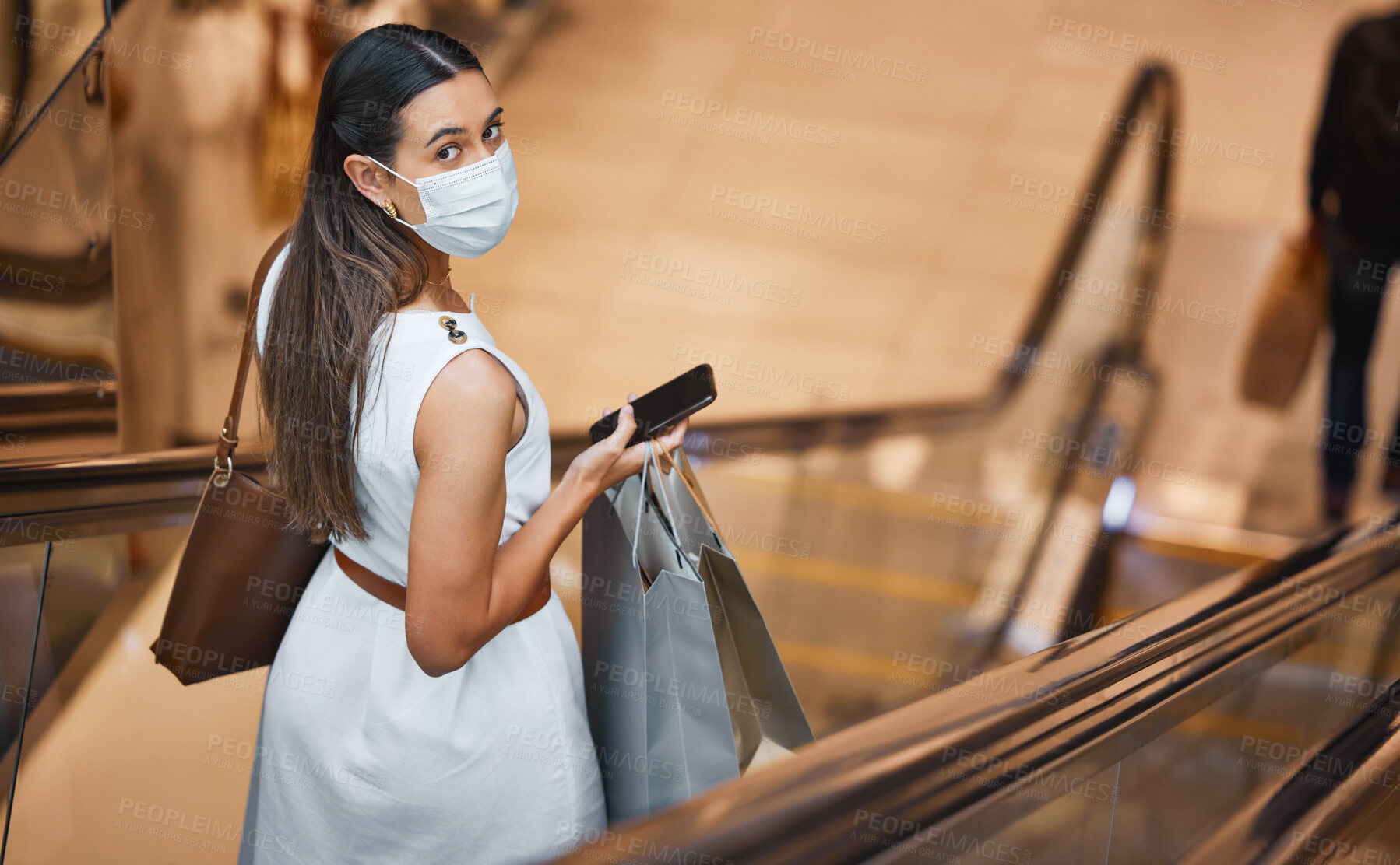 Buy stock photo Mask, portrait and retail with woman in shopping mall, on escalator for bargain, deal or sale. Bags, covid and phone with customer or shopper in store from above for commerce and communication