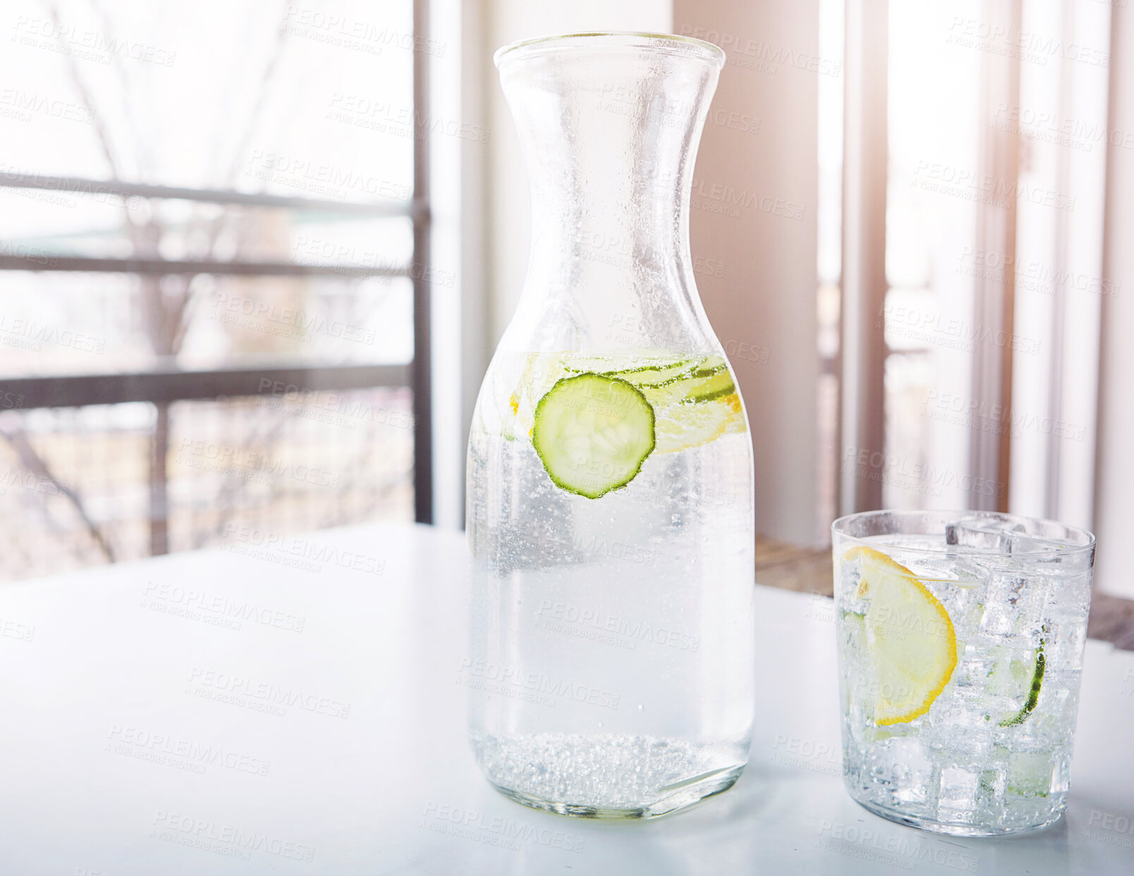 Buy stock photo Glass, jug and lemon water with cucumber on table for detox, nutrition and healthy drink in house. Citrus, fruit and ice with liquid in container for antioxidants, vitamin C and organic hydration