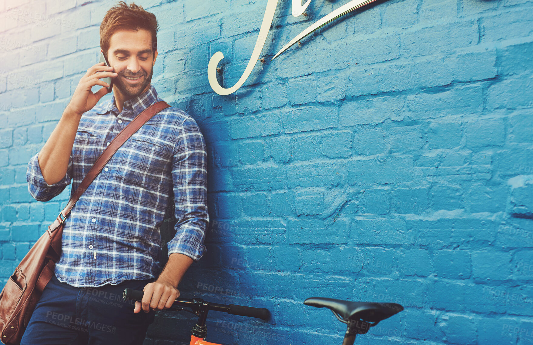 Buy stock photo Talking, phone call and happy man on bicycle in city for eco friendly transportation by brick wall. Mobile, conversation and person with bike for communication, travel and commute by space outdoor