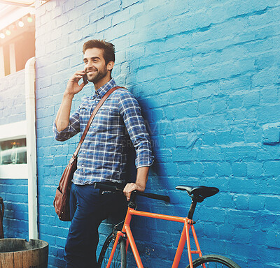 Buy stock photo Conversation, phone call and happy man on bicycle in city for eco friendly transportation. Mobile, talking or person with bike for communication, commute or travel plan by space outdoor by brick wall