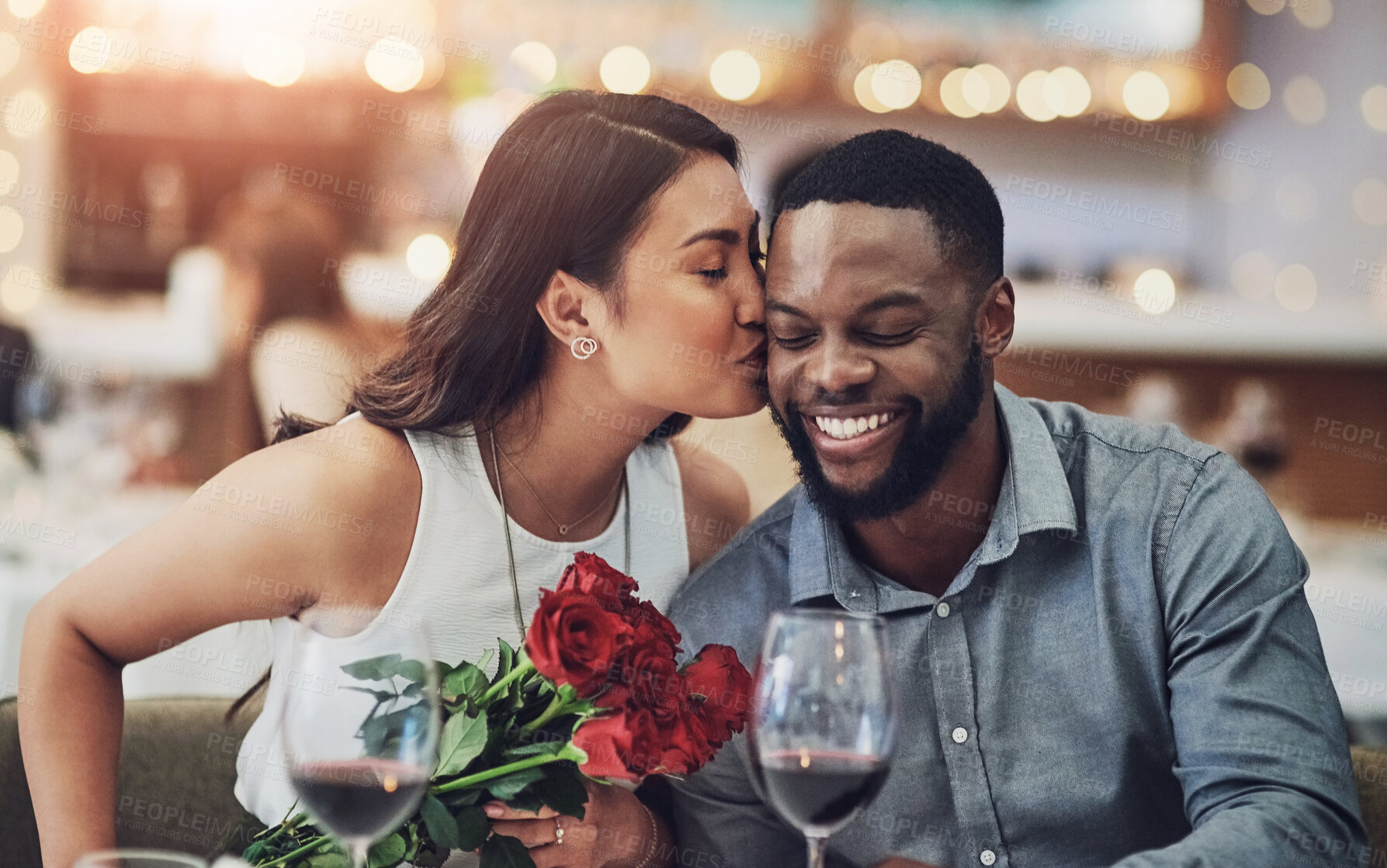 Buy stock photo Couple, dinner and kiss on date in restaurant for flowers, romance or commitment to relationship. Interracial, people and happy with celebration for anniversary, honeymoon or valentines day with wine