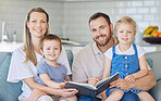 Portrait of young parents with children reading a book, caucasian family of four smiling and enjoying a story. Cheerful couple teaching their two children how to read while sitting on a sofa