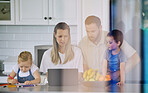 Caucasian family having lunch time with fresh fruit in a bright kitchen. Affectionate father helping his wife while she's using a laptop and sitting with her kids at home while little daughter draws in coloring book