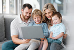 A happy family using a laptop on the sofa with children at home. Smiling man and woman relaxing online with their sons with a computer. Cute little boys sitting and learning  with their parents