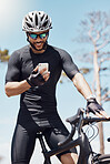 Smiling young male cyclist wearing helmet and glasses while using his smartphone and standing with his bike. Fit sportsman using app or sending a message on mobile phone while cycling outdoors