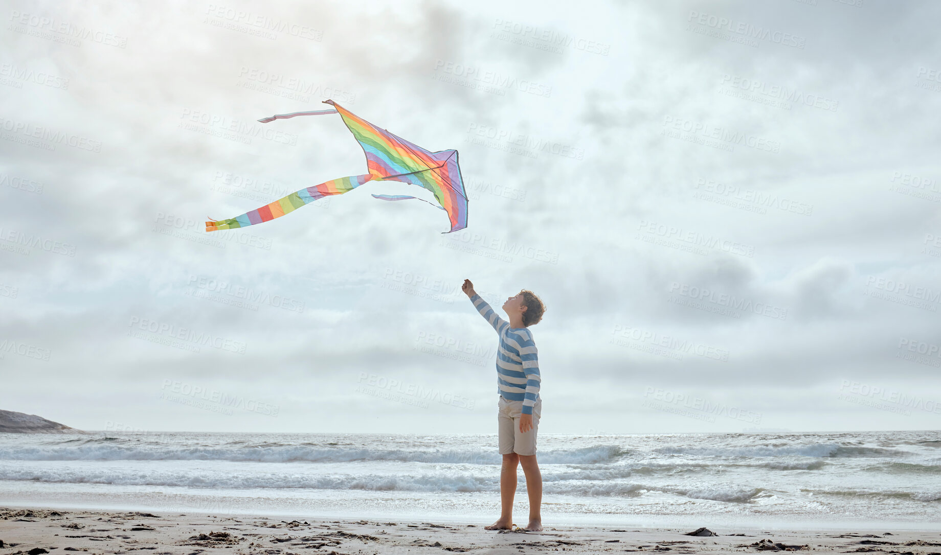 Buy stock photo Boy, child and kite by ocean for vacation, playful and outdoor in wind by waves, clouds and walk at beach. Kid, toys and rainbow fabric for flight, games and holiday by sea with adventure in Spain