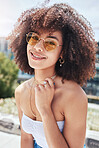 Portrait of young trendy beautiful mixed race woman with an afro smiling and posing alone outside. Hispanic woman wearing sunglasses and feeling happy. Fashionable African American woman in the city