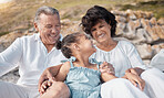 Smiling mixed race grandparents sitting with granddaughter on a beach. Adorable, happy, hispanic girl bonding with grandmother and grandfather outside on weekend. Seniors and child enjoying free time