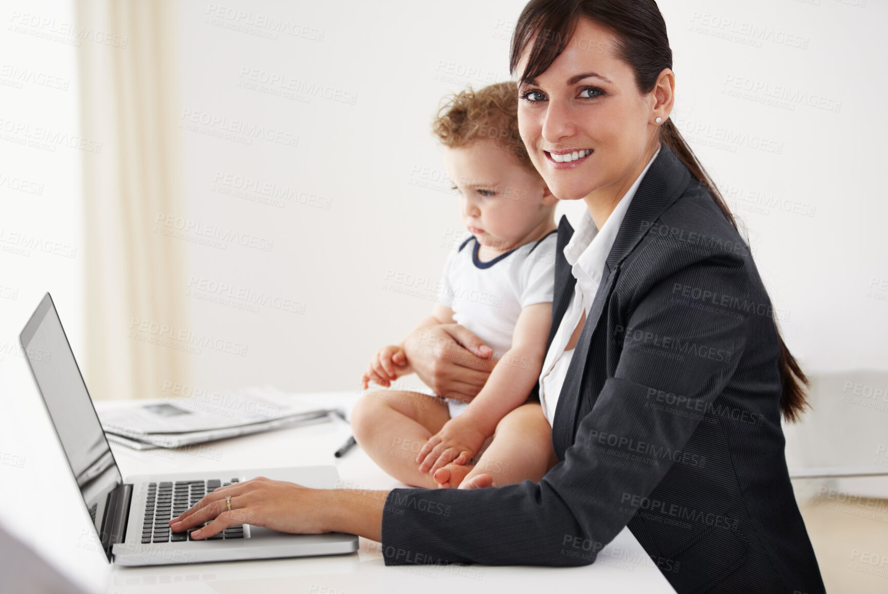 Buy stock photo Young working mom smiling at the camera while holding a camera and typing on her laptop