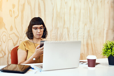 Buy stock photo Shot of a young designer using her cellphone while working on a laptop in an office