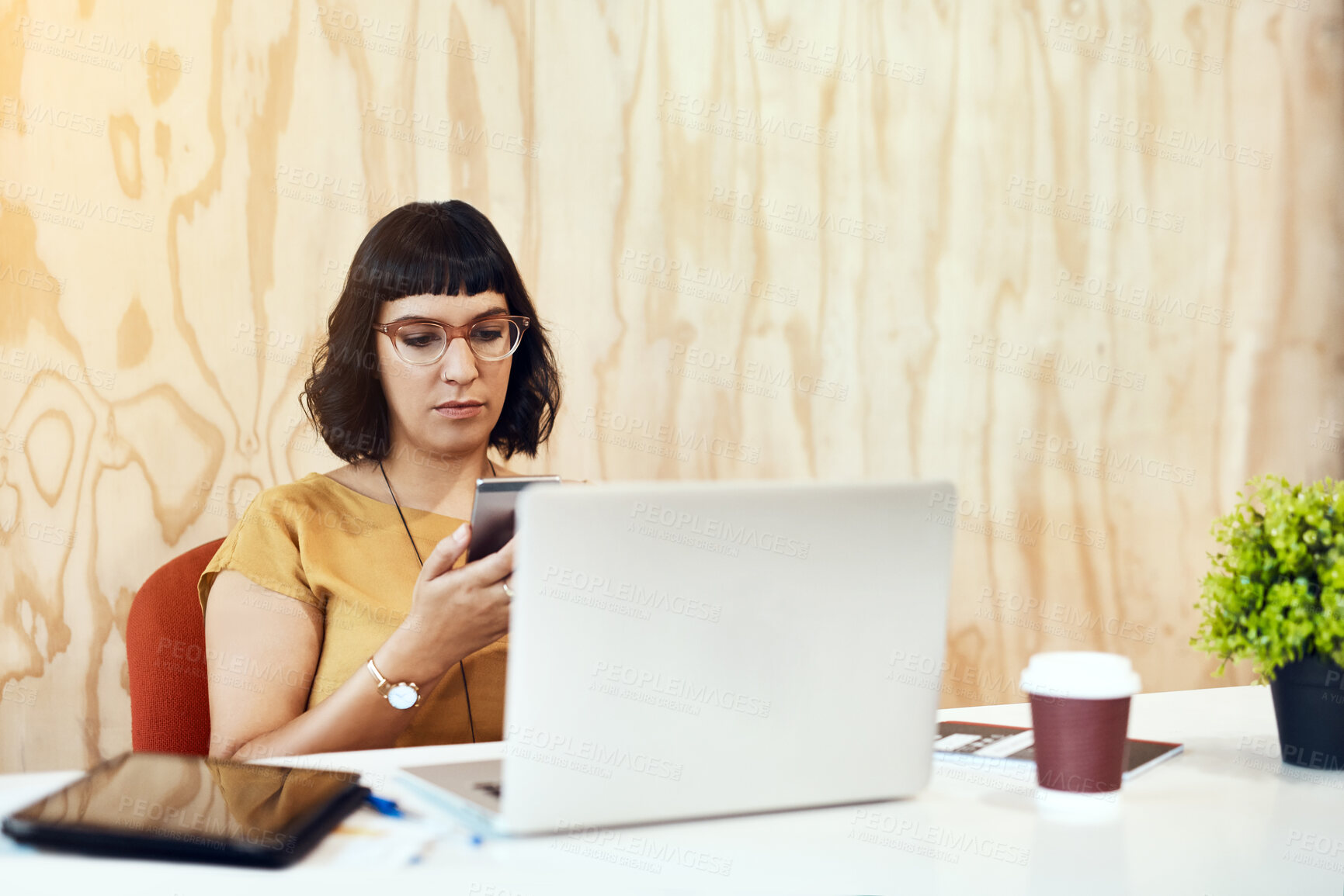 Buy stock photo Shot of a young designer using her cellphone while working on a laptop in an office