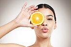 Young happy beautiful mixed race woman pouting while holding an orange and posing against a grey studio background alone. Carefree hispanic female smiling showing a fruit while standing against a background