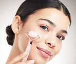 Young happy mixed race woman holding and using a jade roller during her skincare routine while posing against a grey studio background. Carefree hispanic female taking care of her skin against a background