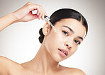 Portrait of a young mixed race woman applying serum to her face while standing against a grey studio background alone. One hispanic female doing her skincare routine while standing against a background