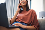 Mixed race woman talking on her mobile phone while using a laptop and working remotely from the comfort of her couch. Young female doing freelance work while talking on the phone and working online