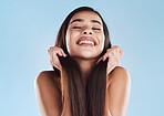 One beautiful young hispanic woman touching her sleek, silky and healthy long hair while smiling against a blue studio background. Confident and happy mixed race model with flawless complexion and natural beauty