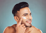 Portrait of one smiling young indian man applying moisturiser lotion to his face while grooming against a blue studio background. Handsome guy using sunscreen with spf for uv protection. Rubbing facial cream on cheek for healthy complexion and clear skin