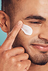 Closeup of one young indian man applying moisturiser lotion to his face while grooming against a blue studio background. Handsome guy using sunscreen with spf for uv protection. Rubbing facial cream on cheek for healthy complexion and clear skin