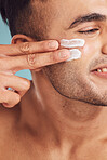 Closeup of one young indian man applying moisturiser lotion to his face while grooming against a blue studio background. Handsome guy using sunscreen with spf for uv protection. Rubbing facial cream on cheek for healthy complexion and clear skin