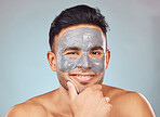 Portrait of one handsome young indian man applying an anti aging facial mask against a blue studio background. Mixed race guy wearing a moisturising clay or charcoal cream product on his face to get rid of blackheads for healthy, smooth and soft skin