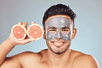 Portrait of one handsome young indian man applying a detoxifying clay or charcoal facial mask while holding grapefruit against a blue studio background. Guy using moisturising products with natural ingredients on his face for healthy, smooth soft skin