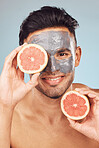 Portrait of one handsome young indian man applying a detoxifying clay or charcoal facial mask while holding grapefruit against a blue studio background. Guy using moisturising products with natural ingredients on his face for healthy, smooth soft skin