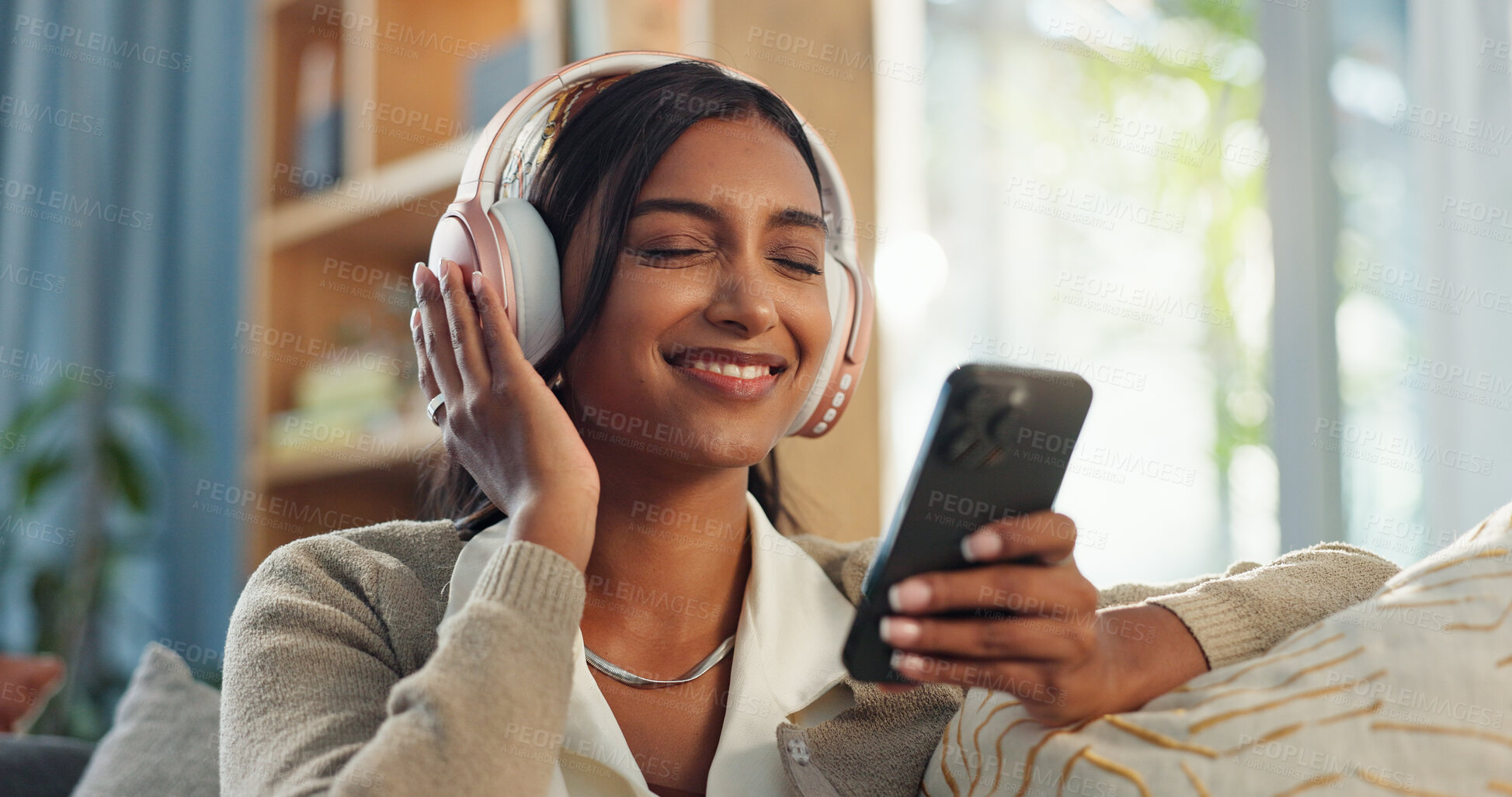 Buy stock photo Headphones, indian woman and cellphone for audio in home, listen and song in living room. Joyful, lounge and online music streaming for sing along app, karaoke and games for female person on sofa 