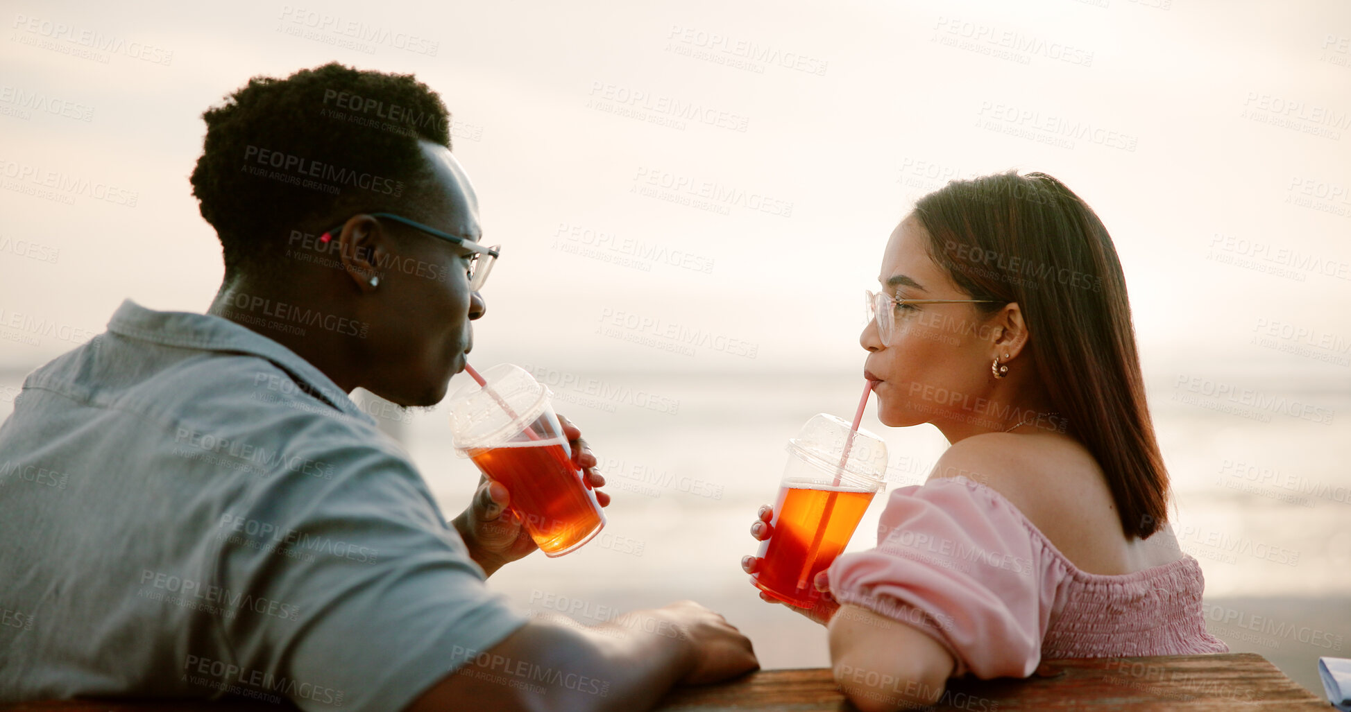 Buy stock photo Couple, drinks and relax on date at beach for bonding, relationship and romance with summer sunset. Interracial people, love and beverage at ocean for support, trust and loyalty on holiday vacation