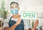 Portrait of one young hispanic man wearing a face mask and hanging up an "open" sign in at a window on display in a cafe or store. Happy mixed race guy excited to open his shop and welcome customers during covid pandemic