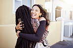 Cheerful young woman holding her smartphone while hugging her friend on sidewalk. Female friends embracing and greeting each other on city street