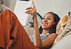 Smiling mixed race woman browsing internet on digital tablet at home. Happy hispanic lying down on living room sofa alone and using technology. Relaxed woman scrolling and searching online on weekend